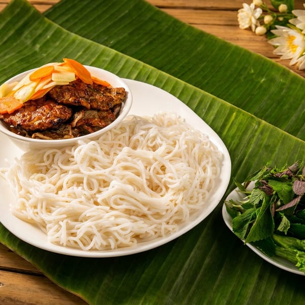 Rice vermicelli noodles served with grilled meat, pickled vegetables, fresh herbs, and lettuce on a banana leaf.