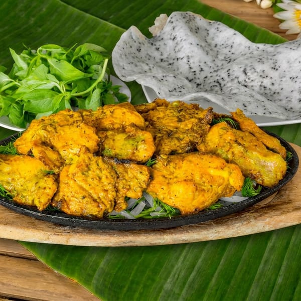 Golden-orange grilled fish pieces served on a sizzling platter with greens, crisp rice crackers, banana leaves, and decorative flowers.