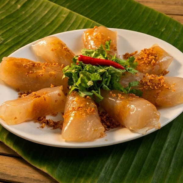 Plate of translucent rice cakes topped with fried garlic, cilantro, and red chili, arranged on a banana leaf over a wooden table.