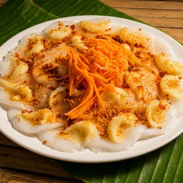 White plate of steamed rice cakes topped with shredded carrots and crispy garlic, arranged on a banana leaf over a wooden table.