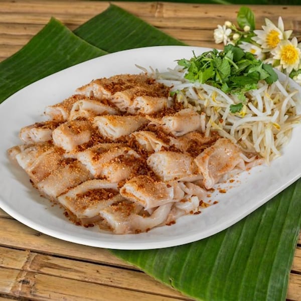 Grilled rice noodle rolls topped with toasted spices, served with bean sprouts, herbs, and flowers on a banana leaf-lined plate.